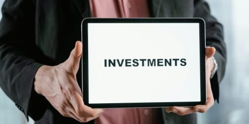 A businessman in a suit holds a tablet displaying the text 'INVESTMENTS'.